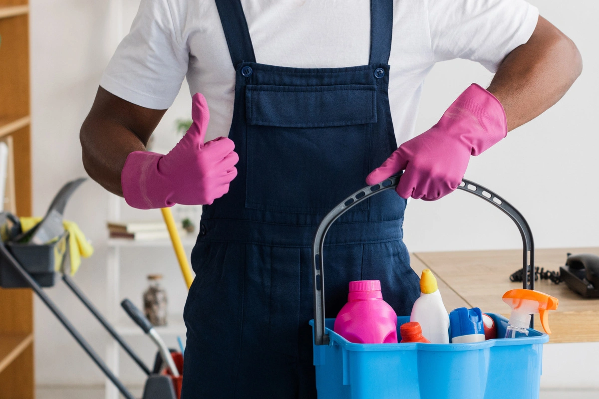 Entreprise de nettoyage : Un agent de nettoyage donne un pouce en l'air, tenant un seau rempli de produits de nettoyage, symbolisant la satisfaction et la qualité des services offerts.